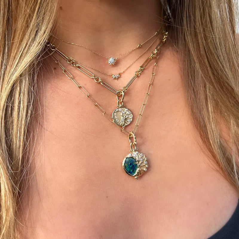 Wearing layered necklaces on a Three Stories Jewelry 14k gold chain with star and circular pendants featured on an alternating paperclip chain of 16 inches, and a vibrant blue-green stone. Styled in a dark top, their long hair cascades over shoulders.