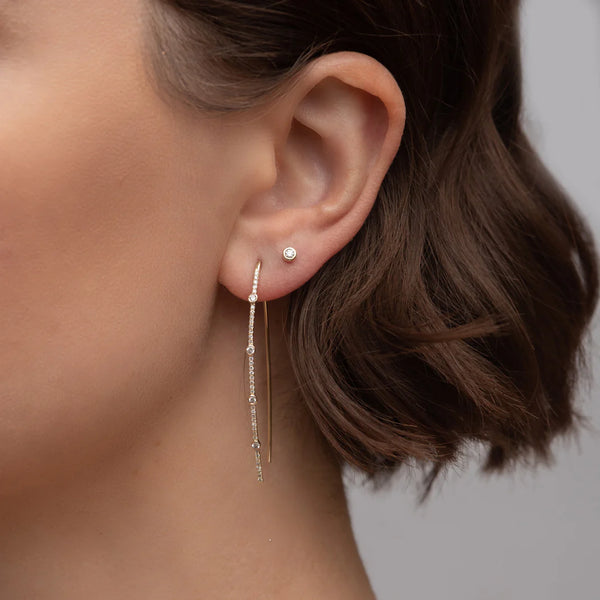 A close-up shows a person with short, dark brown hair wearing two elegant earrings: a small 14k gold stud in the upper lobe and Rachel Reids Mixed Diamond Wire Earrings hanging from the lower lobe, set against a plain gray background.