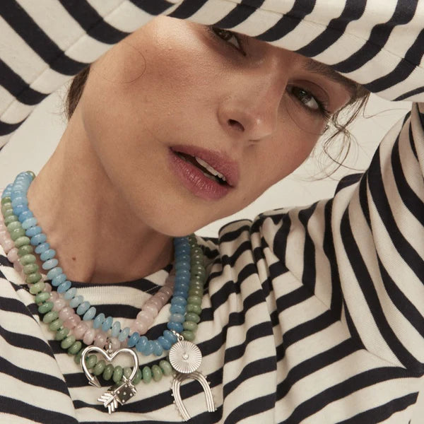 A woman with pink lips poses in a black and white striped top, showcasing the Adina Reyter Spring into Pink Gemstone Necklace featuring colorful beads and large silver heart and sunburst pendants.