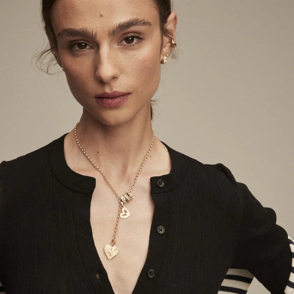 A person with light skin and brown hair, wearing a black top, gold earrings, and the Adina Reyter Rolo Chain Lariat Necklace, stands against a plain background.