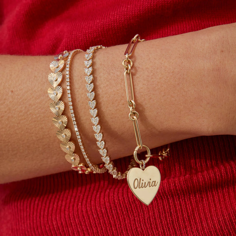 A wrist is adorned with multiple gold bracelets from ef collection, including a dazzling mixed chain enhanced bracelet and two with detailed designs. One chain bracelet features a large heart charm engraved Olivia. The background is red fabric.