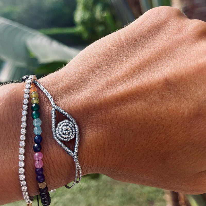 A close-up of a wrist wearing the Oiya Women’s Seven Chakras Bracelet, handmade with multicolored beads and a silver spiral-eye charm. Blurred green foliage appears in the background.