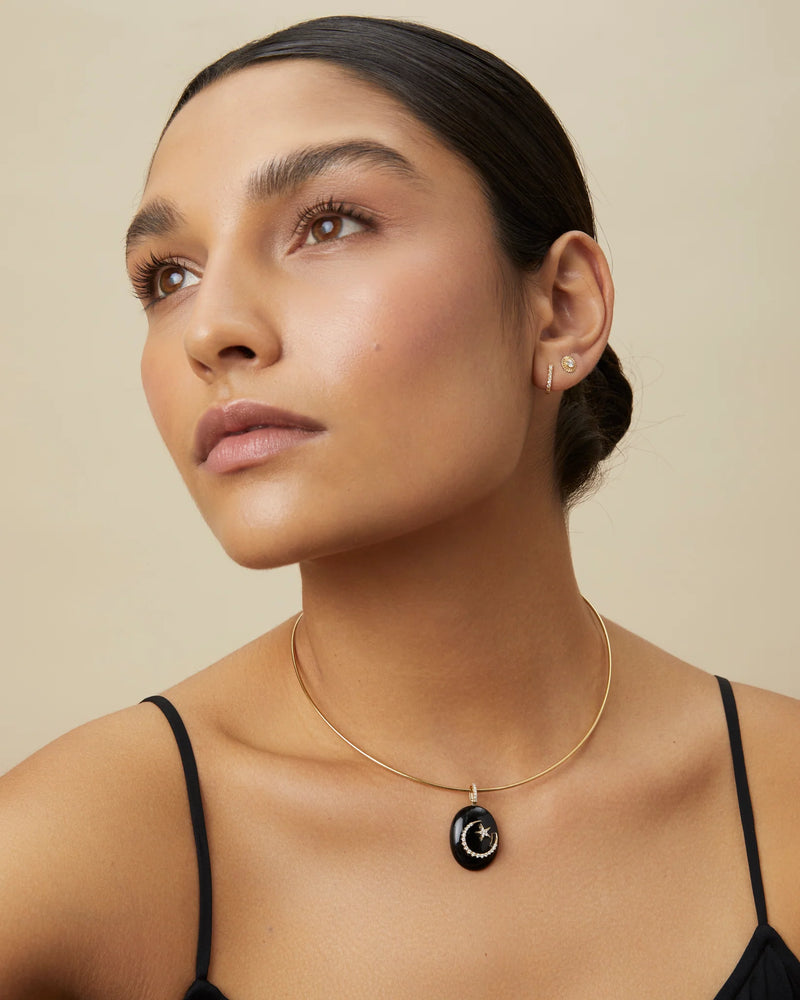 A woman with smooth dark hair pulled back wears the Omega Chain by Kate Collins Jewelry—a thin yellow gold necklace—paired with a black oval pendant, small hoop earring, and a slim strap top, gazing upward against a neutral background.