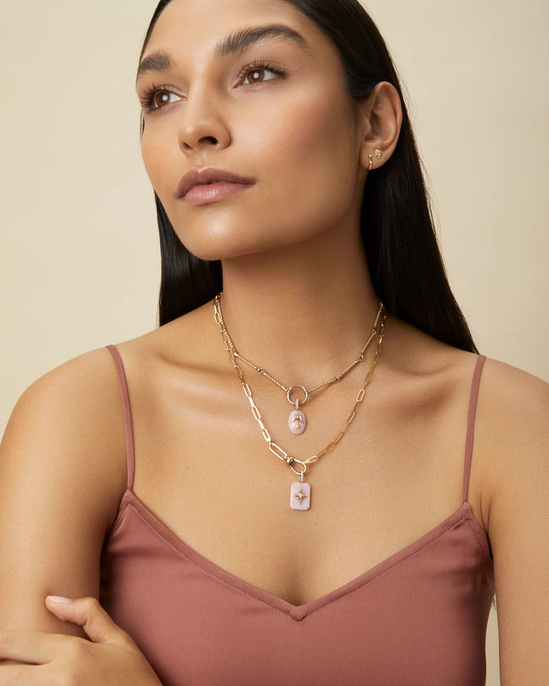 A woman with long dark hair and flawless skin wears a mauve spaghetti strap top and the Kate Collins Jewelry Twisted Bar Necklace. She gazes upward calmly, delicate elegance enhanced against a neutral beige backdrop.