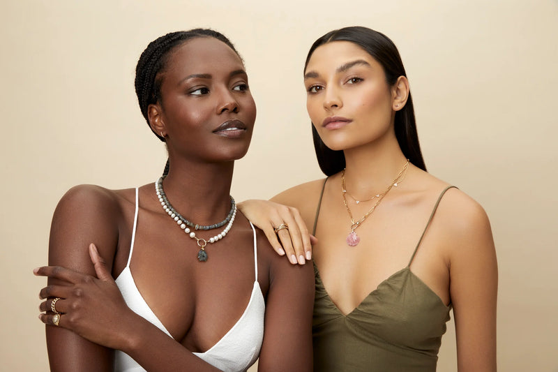 Two women pose against a beige backdrop in white and olive green spaghetti strap tops, both wearing delicate rings and the Pink Rhodochrosite Scallop Shell Pendant by Kate Collins Jewelry, featuring a diamond bail set in 14K gold.