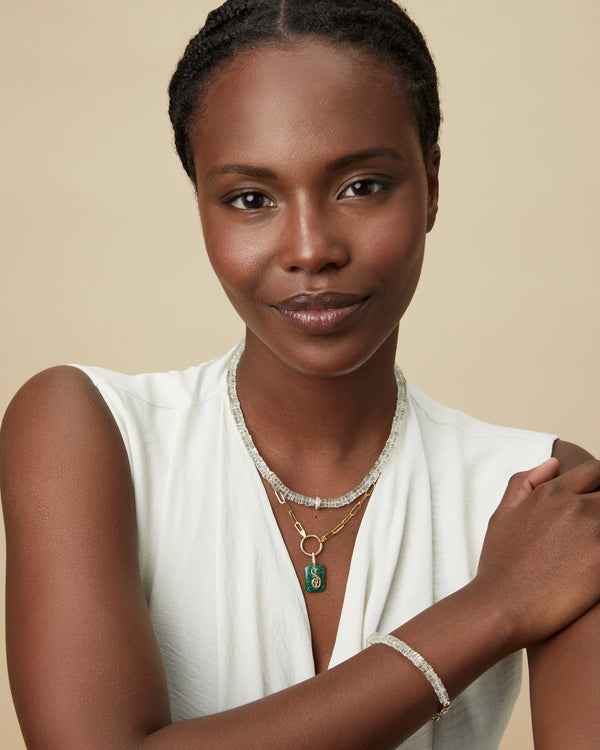 A woman with braided hair smiles at the camera, wearing a white sleeveless top and layered necklaces, including a Kate Collins Jewelry charm with the Small Emerald Cut Cabochon Monogram. Her crossed arms showcase a chic bracelet against the soft beige background.