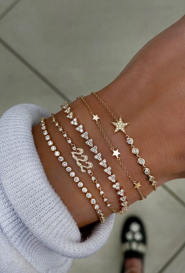 A wrist stacked with five delicate bracelets, including Luna Skye’s 14kt gold full three diamond cluster tennis bracelet; an emerald bracelet with stars, hearts, and gemstones. A white ribbed sleeve and embellished shoe appear in the background.