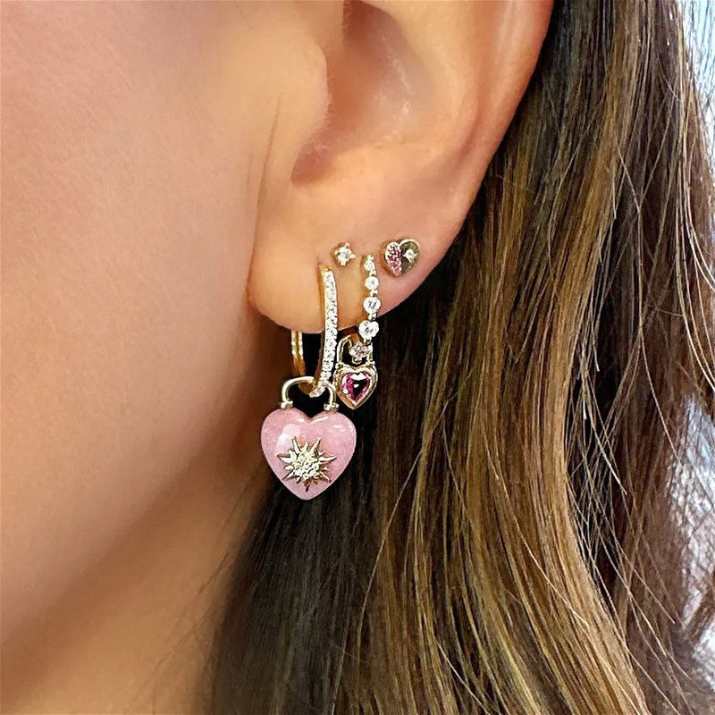A close-up of an ear with multiple piercings, featuring Three Stories Jewelry’s Beating Heart Medium Blue Sapphire Charms and other sparkling heart-shaped studs, a 14K yellow gold hoop, and an emerald charms stud.