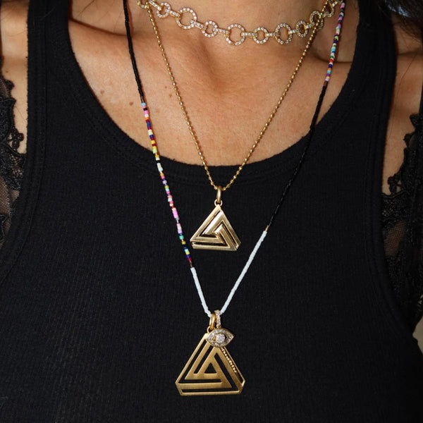 Dressed in a black top, a person models Aaron Bashas Large Abracadabra Triangle Series 4 necklaces: one with an 18K gold-plated triangle pendant, another portraying empowerment with an eye motif and beads, plus a colorful beaded choker.