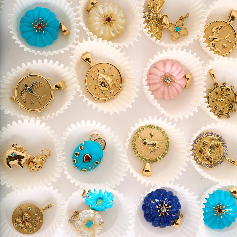 The Jane Win collection features colorful pendants, including the LUCKY Rabbit JW Small Embellished Pendant Coin in sterling silver. Displayed in white cupcake liners, designs include flowers, stars, and animals as lucky symbols in blue, pink, and gold.