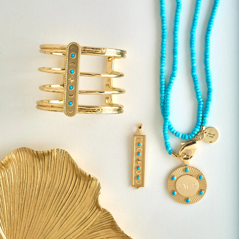 The Jane Win Double Wrap Diamond Cut Beaded Necklace in Turquoise, paired with gold cuff and pendant jewelry, is displayed beside a gold shell dish, featuring blue beads and engraved initials on the round necklace charm.