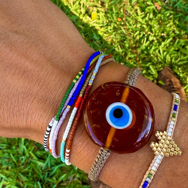 A hand wearing multiple colorful bracelets, including the Trinity Beaded Bracelet by Oiya, with a large brown evil eye charm bracelet, rests on green grass.
