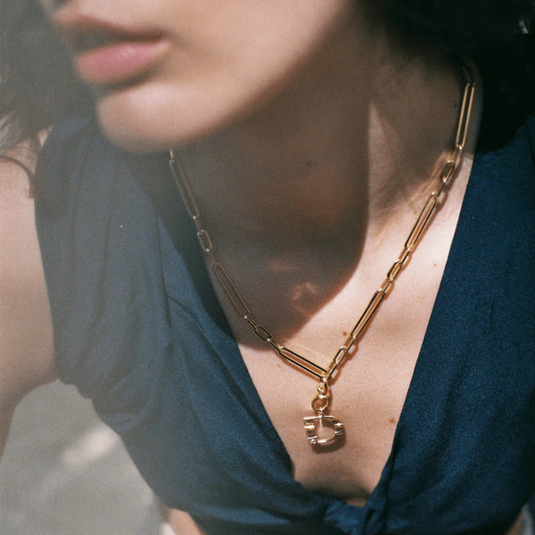 A close-up shows a person wearing zahavas 20 Mika Paperclip Chain Necklace, featuring oval and round links with a padlock pendant. Made of 14k gold, the necklace gleams against their dark blue top in soft, natural lighting.