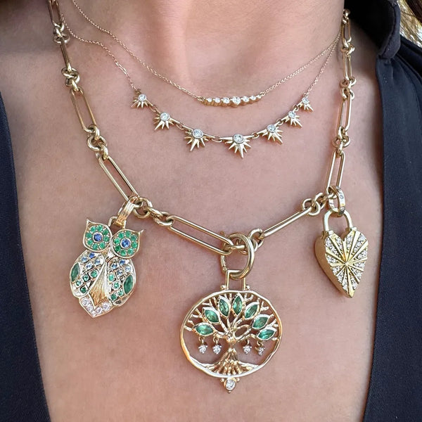 A close-up of a woman's neck adorned with 14K gold jewelry, featuring layered necklaces: star charm chain, chunky owl chain, Three Stories Jewelry's Large Open Tree Of Life Pendant with green leaves, and a heart-shaped pendant.