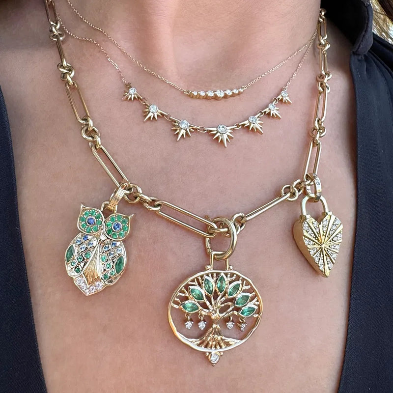 A close-up of a woman's neck adorned with 14K gold jewelry, featuring layered necklaces: star charm chain, chunky owl chain, Three Stories Jewelry's Large Open Tree Of Life Pendant with green leaves, and a heart-shaped pendant.