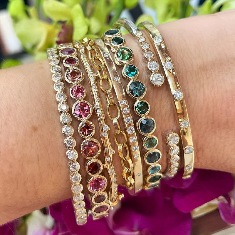 Close-up of a wrist wearing Three Stories Jewelrys Blended Hammered Diamond Bezel Bangle. The 14K yellow gold bangle features colorful gemstones in reds and greens with circular and linked designs, set against a background of blurred green leaves and purple flowers.