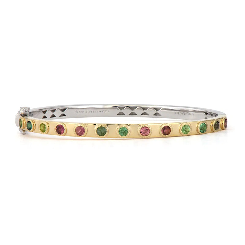 The Two-Toned Flip Colored Tourmaline Bangle by Three Stories Jewelry is a slim bangle with a gold exterior and sterling silver interior, adorned with alternating green and pink tourmalines and featuring a larger clear gemstone at the clasp.