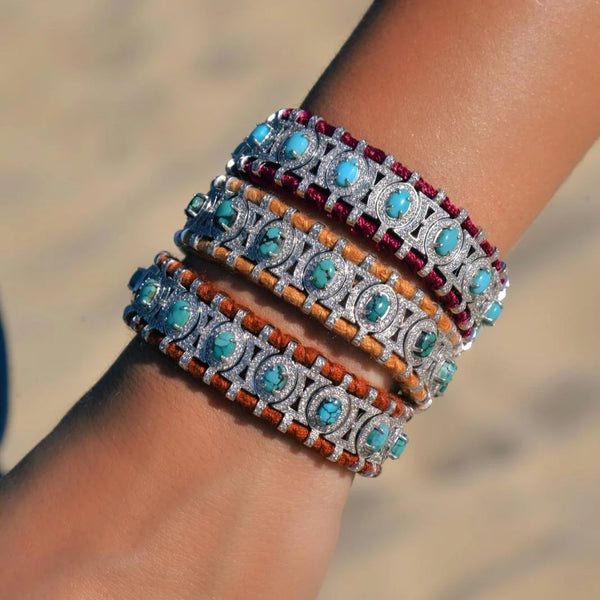 A wrist adorned with four Van Den Abeele Sao Paulo Turquoise bracelets, each featuring turquoise stones in ornate silver frames on red, orange, tan, and brown bands. The vibrant collection contrasts against a soft, blurred sandy background.