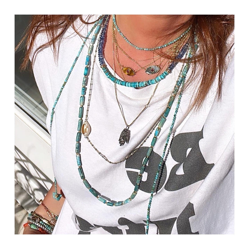 A person dons the KEVIN Lariat from Bohemian Rhapsodie Paris, featuring layered necklaces in gold-plated and turquoise hues with pendants like an evil eye and a Hamsa hand, paired with a white t-shirt showing partially visible black text.