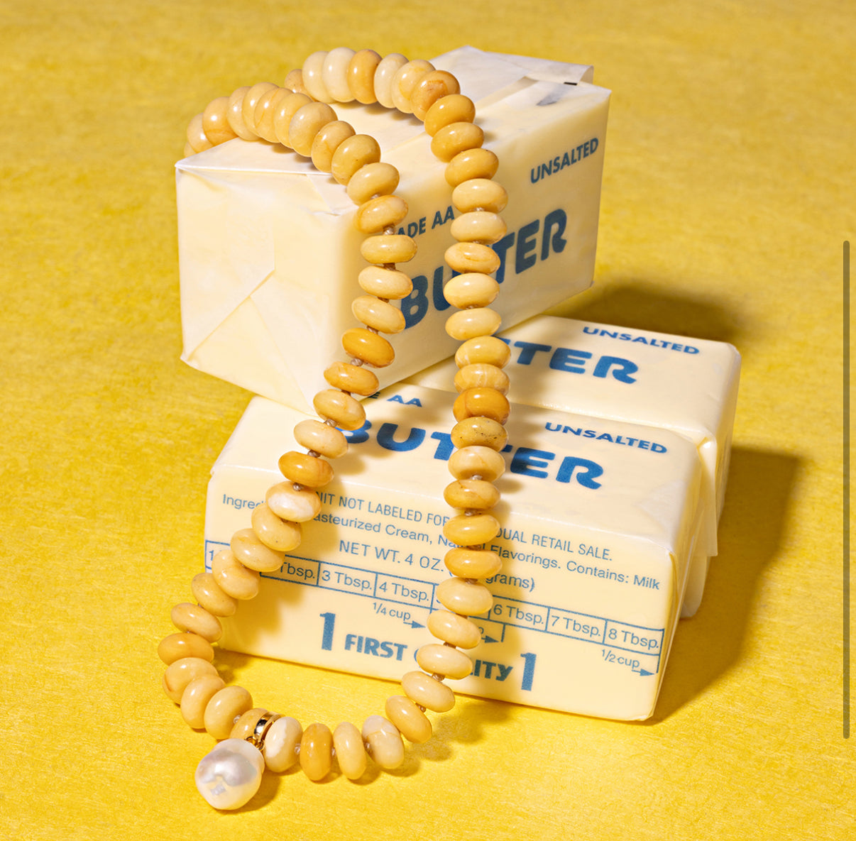 The Adina Reyter Yellow Jade Gemstone Necklace is displayed atop three wrapped sticks of unsalted butter, set against a vibrant yellow background.