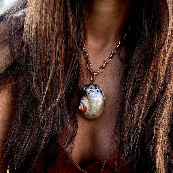 A person with long brown hair wears the Oiya Tori Shell Gold Link Necklace—a handmade piece featuring a large shiny shell pendant on a chunky chain with a gold-plated clasp—over a brown top, centered on their chest.