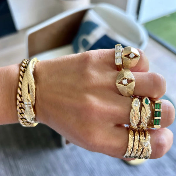 A hand adorned with Helena Rose’s Sophia Serpent Ring featuring an emerald, intricately detailed in 14k yellow gold with a serpent motif, alongside ornate gold rings and a chunky bracelet, set against a blurred indoor background.