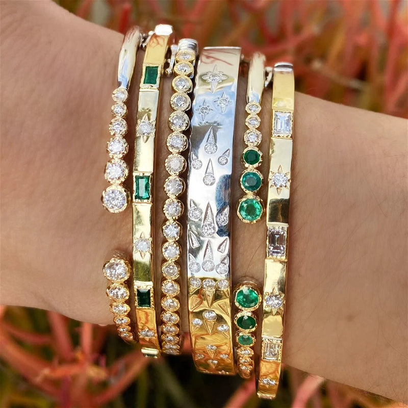 A wrist adorned with six Blended Hammered Diamond Bezel Bangles by Three Stories Jewelry in 14K yellow gold and sterling silver, each boasting exquisite designs with diamonds and emeralds, featuring round, square, and teardrop-shaped gems against a blurred outdoor backdrop.