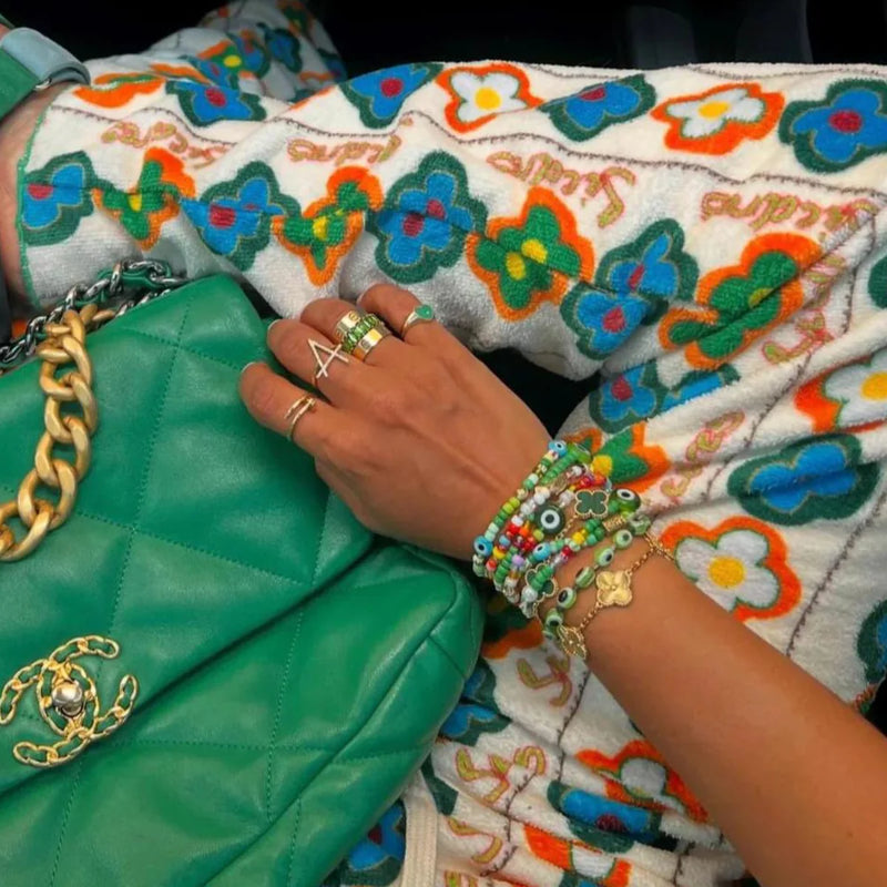 Sitting cross-legged, they showcase floral pants and matching Aaron Basha Womens Blue Beaded Stretch Bracelets. They hold a green quilted bag with a gold chain, and their fingers gleam with rings and diamond-encrusted charms.