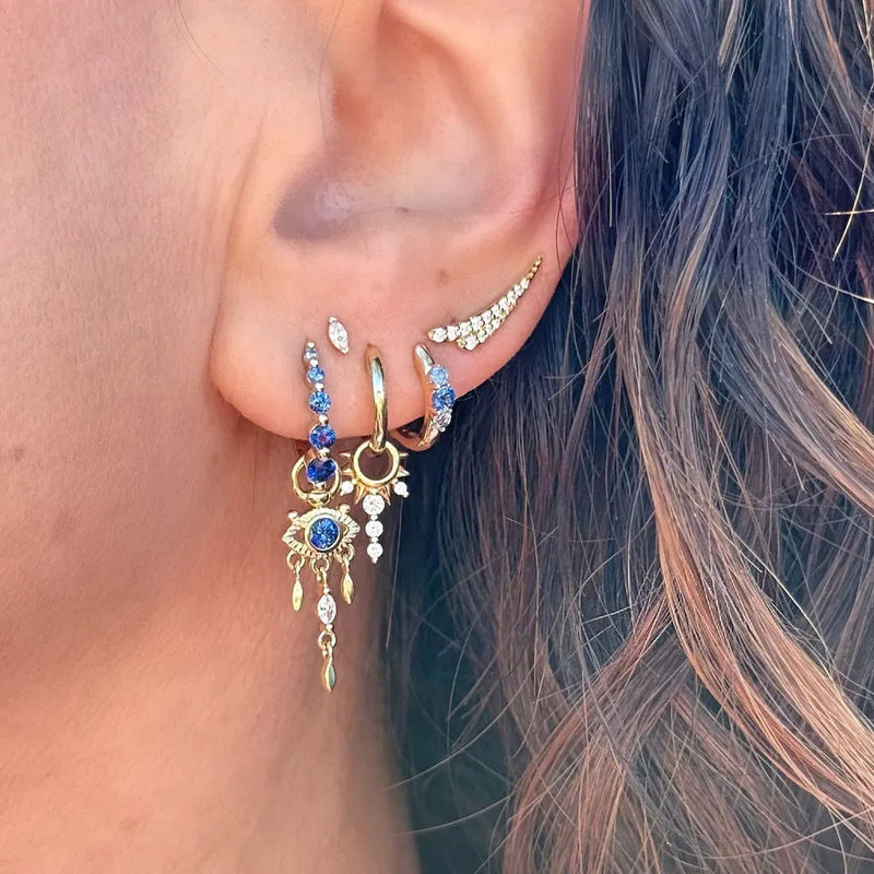 A close-up of an ear with multiple 14K yellow gold earrings, including hoops and studs with blue and white gemstones, plus a dangling Open Evil Eye Sapphire Charm by Three Stories Jewelry. Wavy brown hair partially frames the ear.