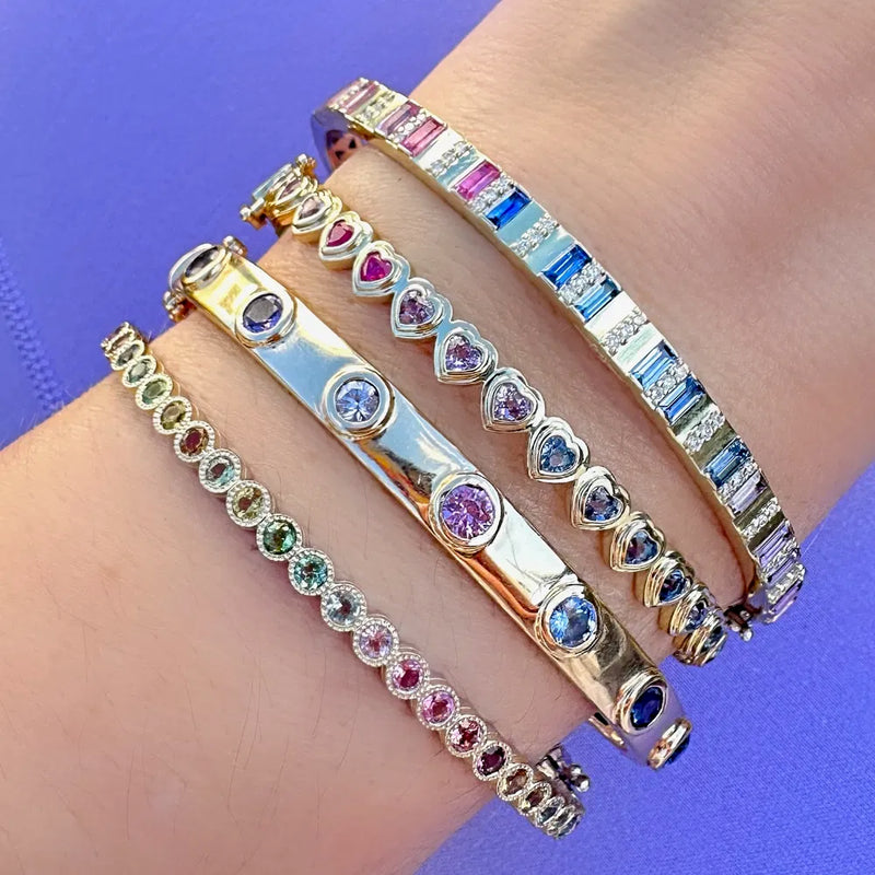 A wrist embellished with four vibrant bracelets shines against a purple backdrop, including a Three Stories Jewelry Hammered Tourmaline Bezel Bangle, beautifully paired with heart-shaped and gemstone-adorned bangles in pink, green, blue, and clear tones.