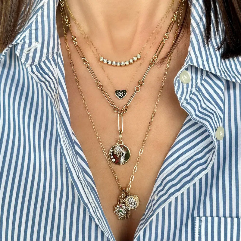 A woman wears a blue and white striped shirt, highlighted by four 14k gold necklaces from Three Stories Jewelry layered around her neck, including an alternating paperclip chain 16 with pendants such as a heart, crescent adorned with stones, and unique charms.