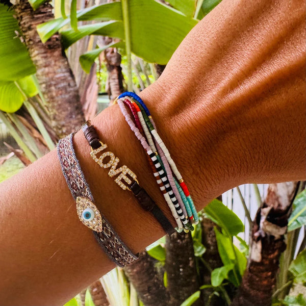 A tan-skinned wrist wears Oiya’s Thea Beaded Bracelet alongside a brown adjustable bracelet with LOVE in gold letters, set against a backdrop of lush green tropical leaves.