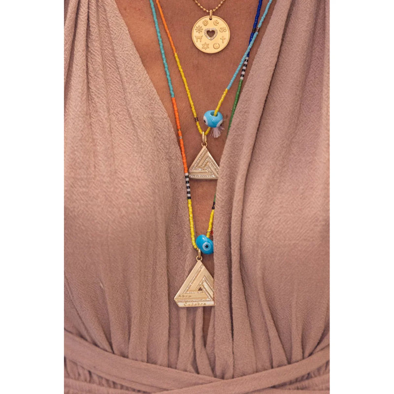 A person in a taupe wrap dress wears an Aaron Basha gold-plated ABRACADABRA pendant symbolizing empowerment and two colorful beaded necklaces from the Large Abracadabra Triangle Series 5, featuring triangular pendants, blue beads, and a small feather.