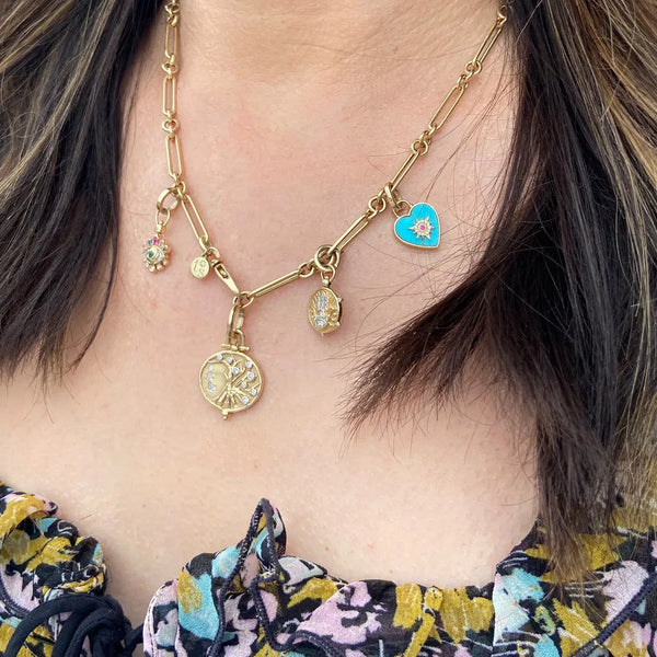 A person dons the stunning Three Stories Jewelrys Alternating Paperclip Chain 16, a 14k gold necklace with paperclip links and charms, like a heart and intricate designs, perfectly matched with a floral top featuring a ruffled neckline.