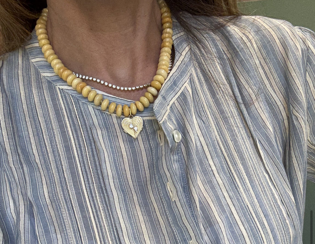 A person wearing a blue and white striped blouse, an Adina Reyter Yellow Jade Gemstone Necklace with a gold heart pendant and hinged charms, and a delicate pearl necklace.