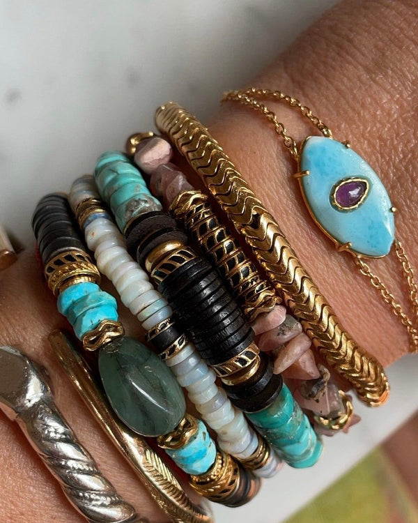 The wrist showcases the unique AYIN Bracelet Larimar from Bohemian Rhapsodie Paris, featuring a gold chain and captivating turquoise stone. Complementary bracelets in black, turquoise, and gold beads enhance the look, while a silver ring on the index finger adds finesse.