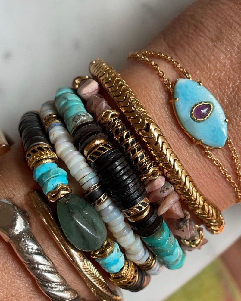 The wrist showcases the unique AYIN Bracelet Larimar from Bohemian Rhapsodie Paris, featuring a gold chain and captivating turquoise stone. Complementary bracelets in black, turquoise, and gold beads enhance the look, while a silver ring on the index finger adds finesse.