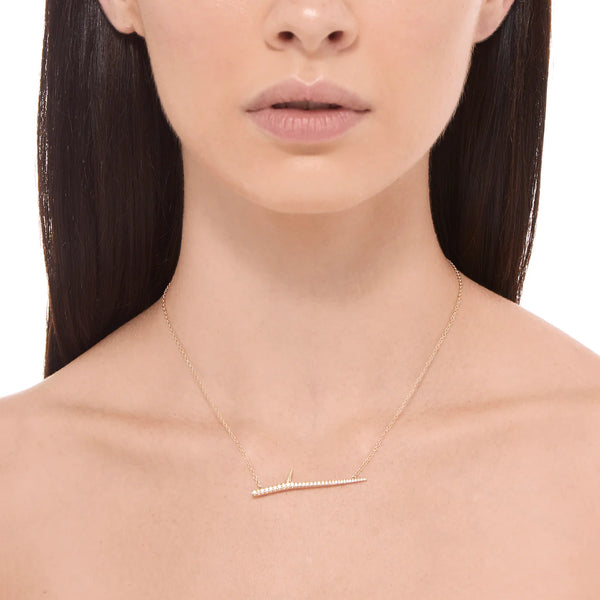 A woman with long, dark hair wears an Elisabeth Bell Thorn Necklace, a delicate gold piece with a sleek horizontal bar adorned with small diamonds. Handmade in Los Angeles, its elegant design beautifully highlights her light skin as it rests against her collarbone.