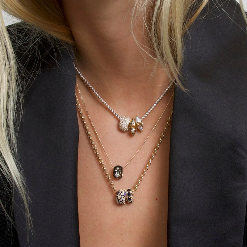A close-up of a person in a black blazer highlights three layered 14k gold necklaces with sparkling round and flower-shaped pendants, along with the Adina Reyter Zodiac Ceramic + Diamond Scorpio Big Bead charm peeking through their blonde hair.