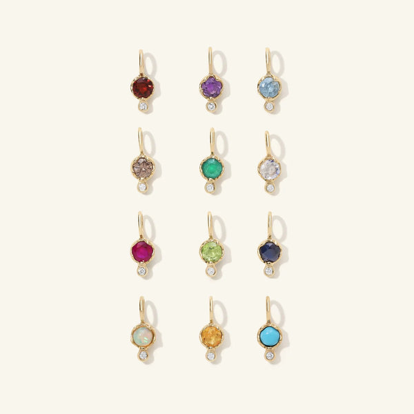 The Birthstone Amulets by zahava feature twelve 10k gold earrings, elegantly displayed in four rows of three. With vibrant red, purple, blue, and green hues, each piece presents a small round gem beneath a larger gemstone pendant.