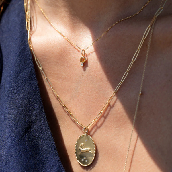 A person wearing layered zahava Birthstone Amulets in 10k gold: one with an oval pendant featuring a small illustration, and the other with a delicate chain and tiny birthstone gemstone, paired with a dark blue top.