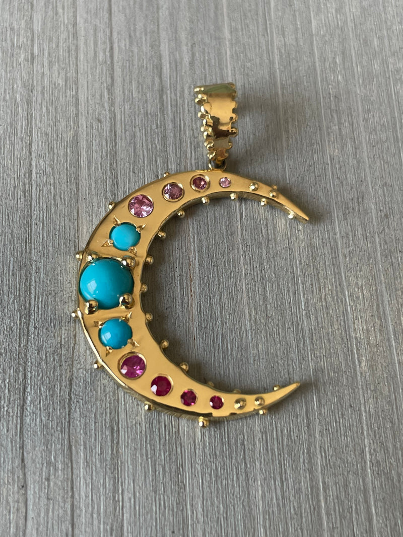 The Boheme Grande Lune Crescent Charm by Delphine Leymarie, crafted from luxurious 18k recycled gold and adorned with pink sapphires and turquoise, shines atop a light gray wooden surface.