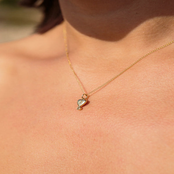 A close-up shows a person wearing zahavas Diamond Byrdie Heart Charm, a delicate gold necklace with a small abstract heart pendant. Light blue and white enamel add detail, while the softly blurred background highlights the jewelry against the skin.