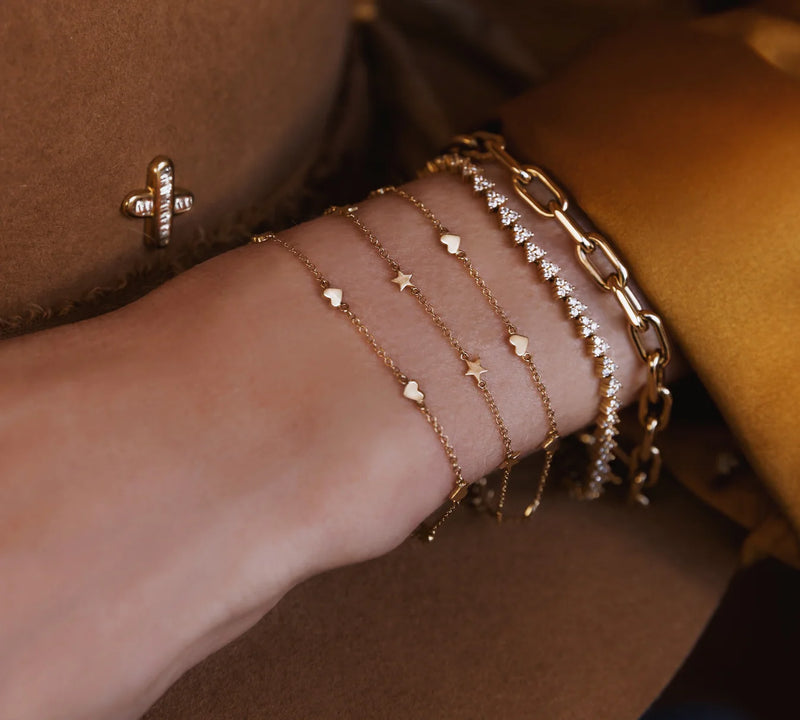A wrist is styled with several gold bracelets, including Luna Skyes 14kt gold full three diamond cluster tennis bracelet, layered with delicate heart and star chains, a chunky gold chain, and paired with a gold buttoned sleeve.