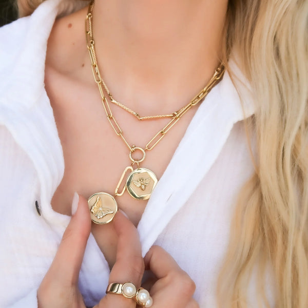 A person in a white top wears celestial layered necklaces: one with a bee pendant and another featuring a 14K Gold Estrella Charm by Kate Collins Jewelry, while holding them with pearl ring-adorned hand. Light blonde hair is partially visible.