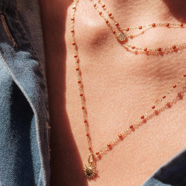 A close-up of a person wearing two delicate Gigi Clozeau necklaces, including the Sun Pendant and small beaded chains, over bare skin with a blue denim shirt.
