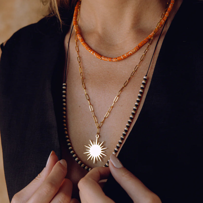 Wearing a black V-neck top, a person layers three necklaces: an orange beaded choker, the Oiya Leilani Sun Chain Necklace, and a black-and-white beaded strand. They hold one strand in their hand.