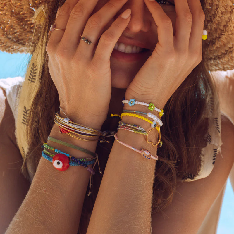 A smiling woman in a straw hat covers part of her face with her hands, showcasing Oiya’s Women’s Seven Chakras Bracelet against a bright, sunny background.