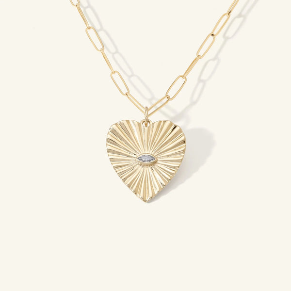 The Diamond Sunbeam Heart Charm by Zahava features a gold heart-shaped pendant with a radiant sunbeam texture and a small diamond marquise stone in the center, hanging from an elongated link gold chain, set against a plain light background.