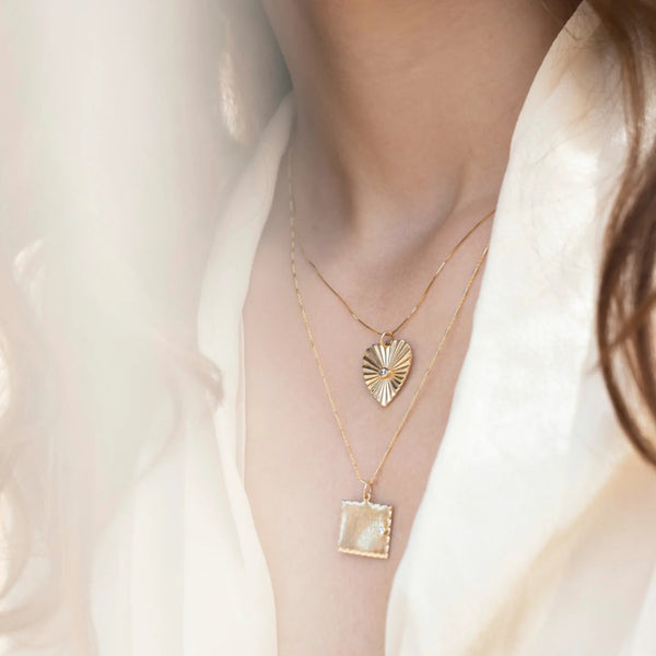 A woman is wearing a cream-colored blouse with two layered necklaces—a zahava Diamond Sunbeam Heart Charm neckpiece above and a square pendant with a pearly finish below—highlighted by sunlight. Her wavy hair partially frames the scene.